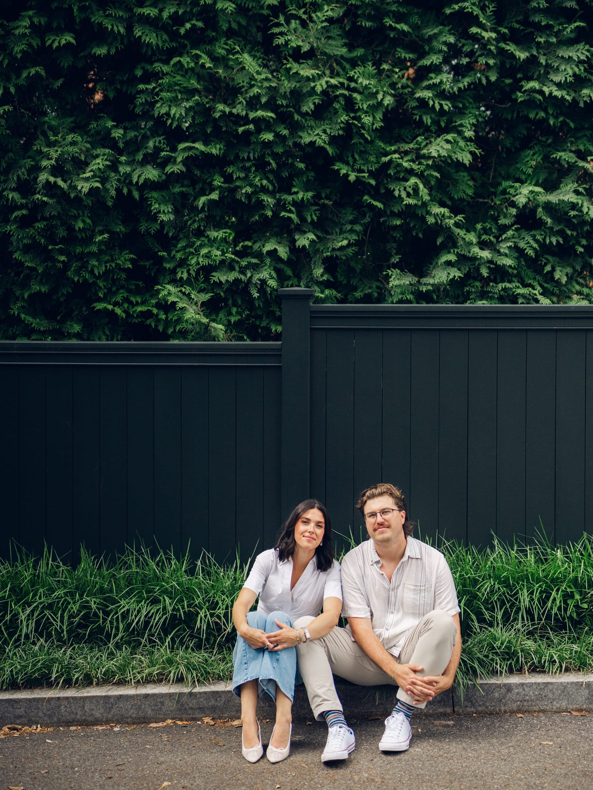 Haley and Slade sitting together on a curb in front of a dark fence and hedge